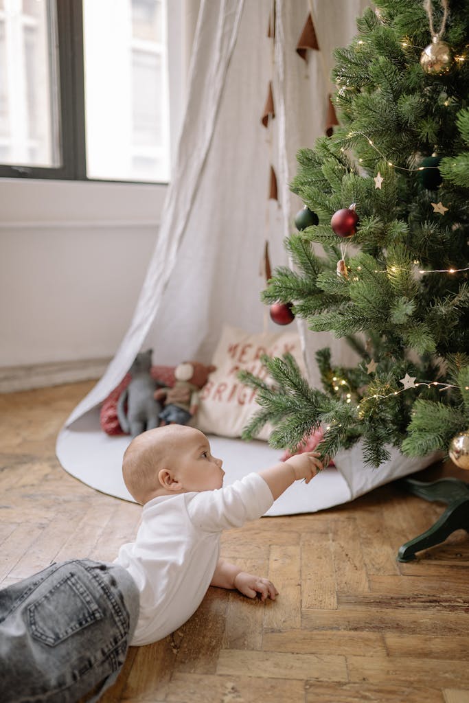 A baby on a wooden floor reaching out to a decorated Christmas tree indoors.