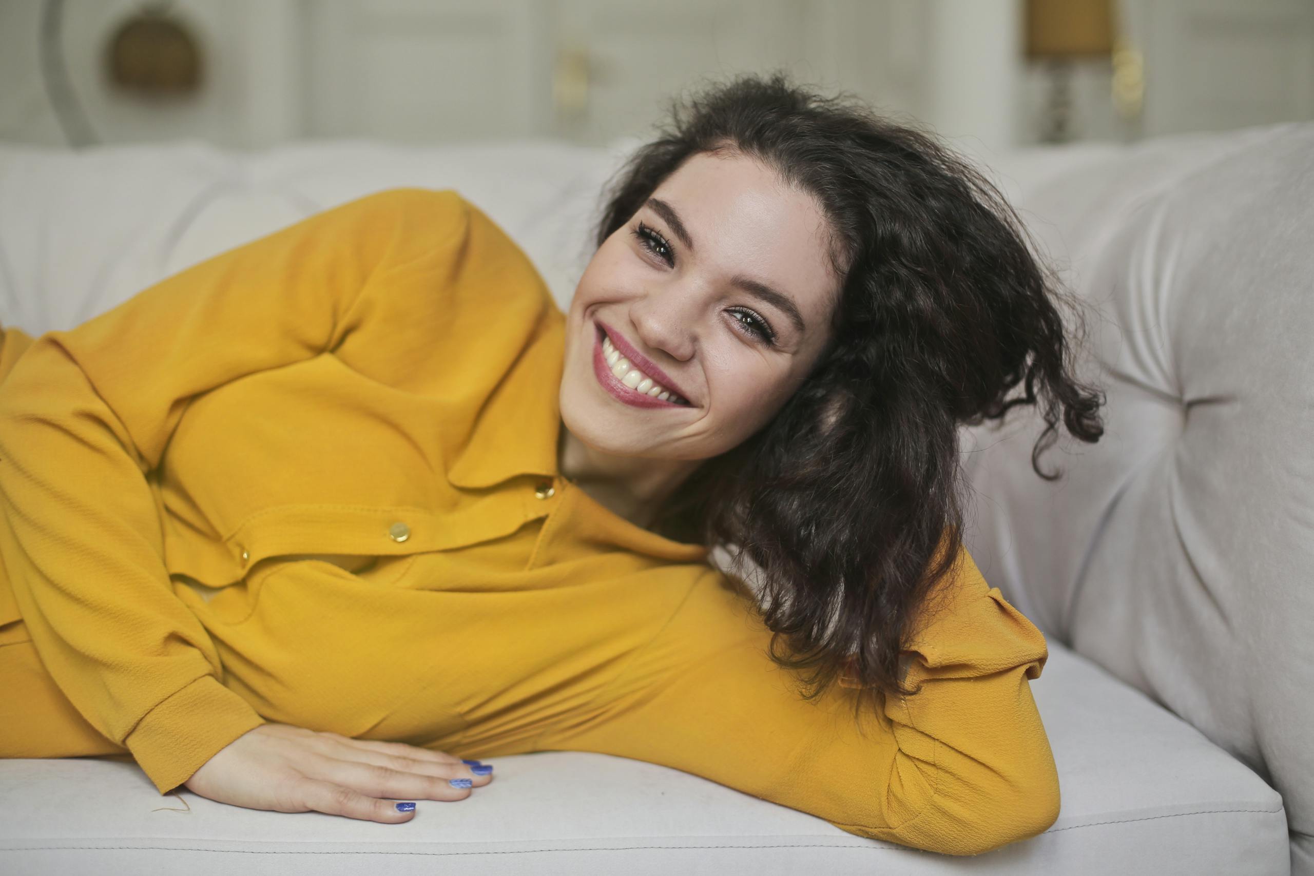 A joyful woman in a yellow outfit smiling while lying on a sofa at home.