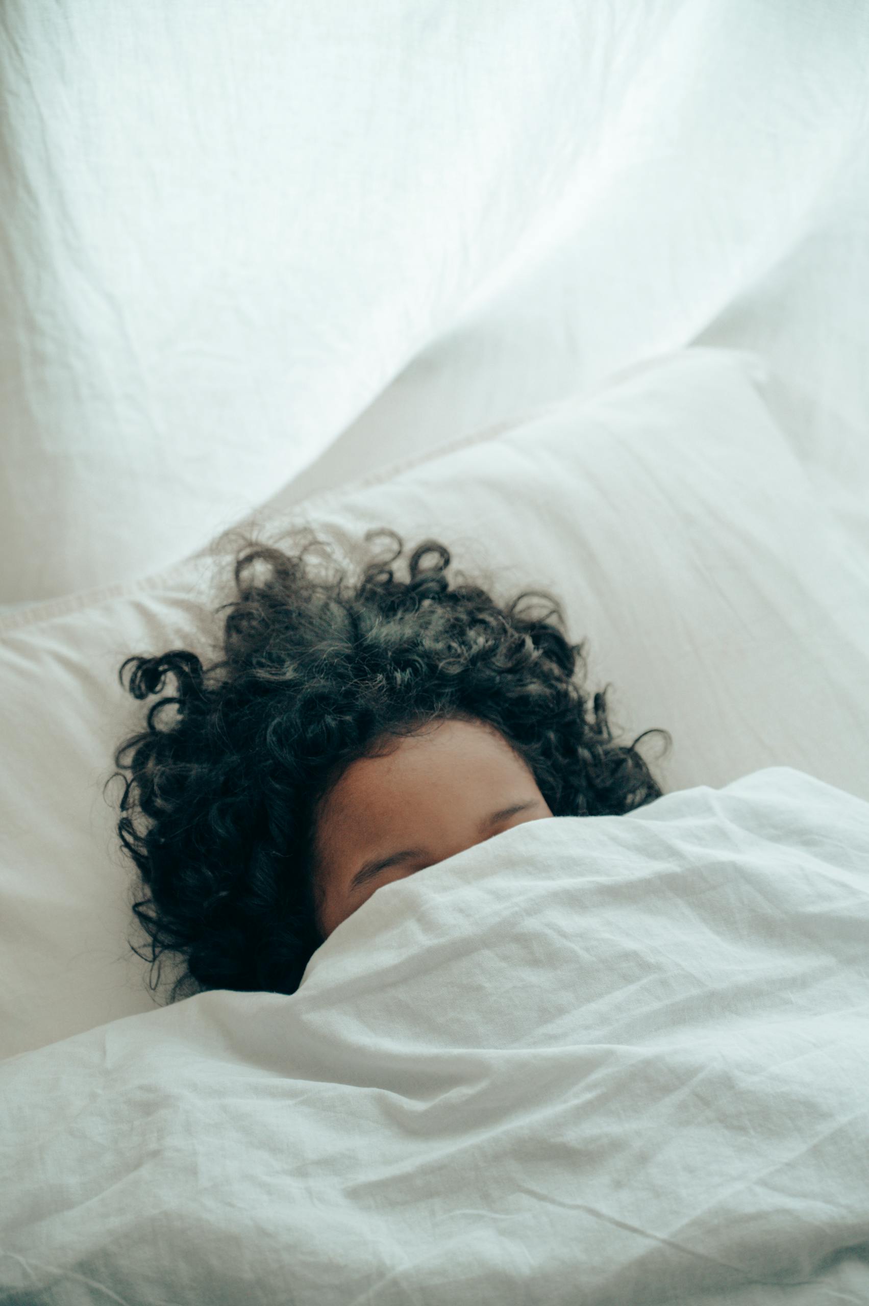 A person with curly hair sleeping peacefully under a white blanket, creating a sense of calm.