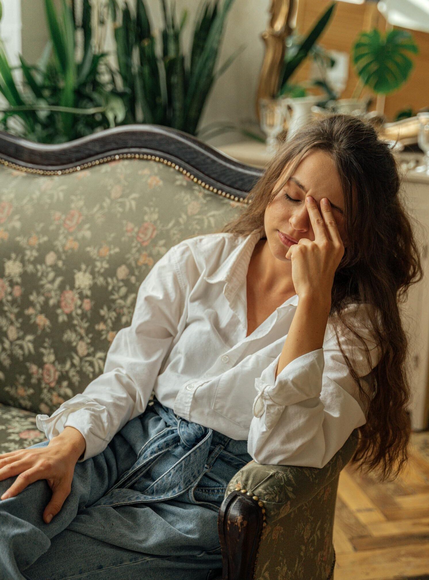 A woman relaxes on a floral vintage sofa in a cozy, light-filled room with plants.