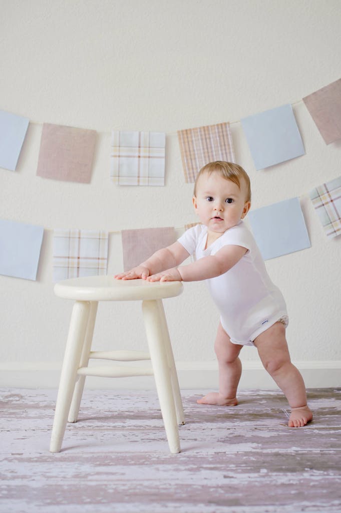 Cute baby standing by a stool indoors with a playful background.