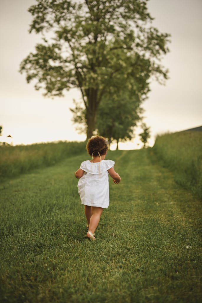 A young child runs freely down a grassy path surrounded by lush trees in a park during summer.