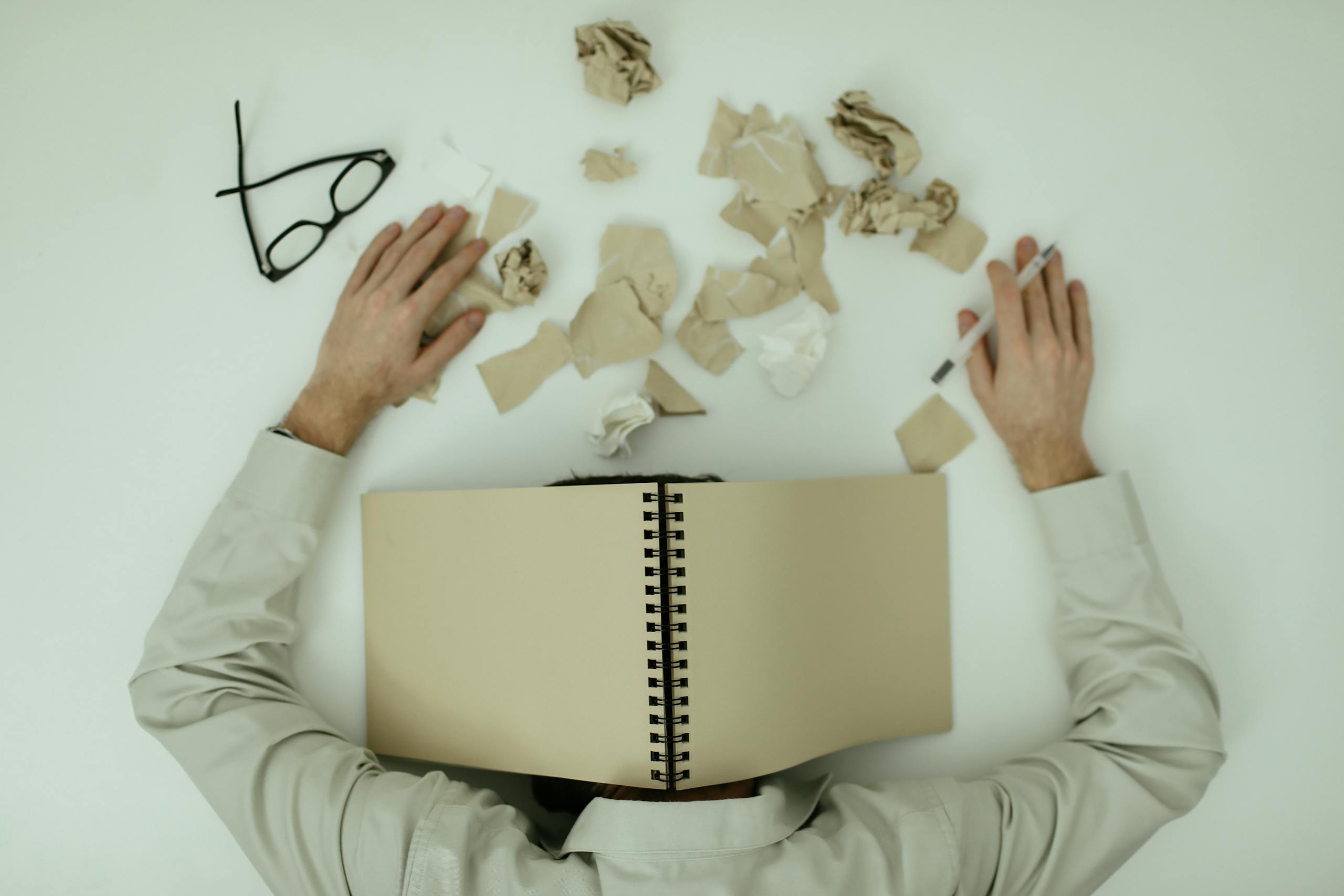 Overhead view of an exhausted man resting with a notepad over his head and crumpled papers around.